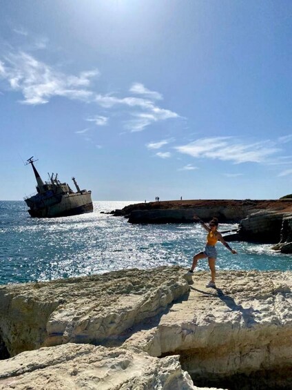A person standing on a rocky outcrop by the sea with a shipwreck in the background. 3