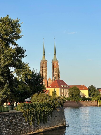 A historic church with two tall spires, a river, and a stone wall with greenery. 2