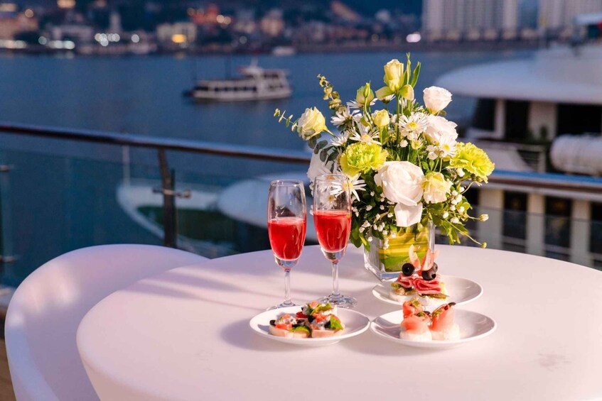 A table with two glasses of red beverage, a floral arrangement, and small plates of food on a balcony overlooking a body of water with a boat in the distance. 2
