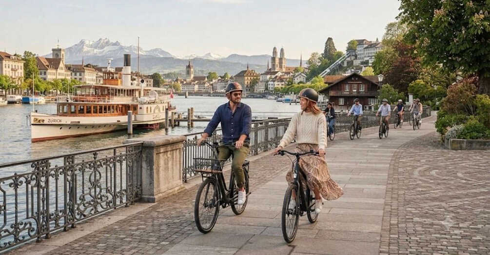 Two people cycling on a paved path by a river with boats and buildings in the background. 5
