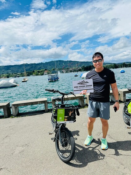 A person standing next to a bicycle with a sign, a body of water, boats, and a hilly landscape in the background. 2