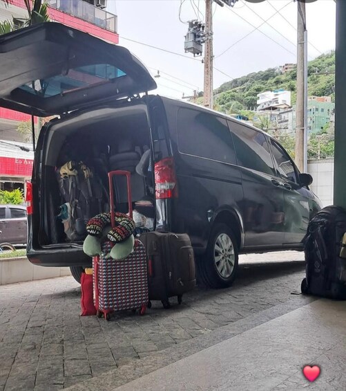 A black van parked on a paved area with its trunk open, revealing a stuffed toy, a patterned suitcase, and a brown suitcase. 2