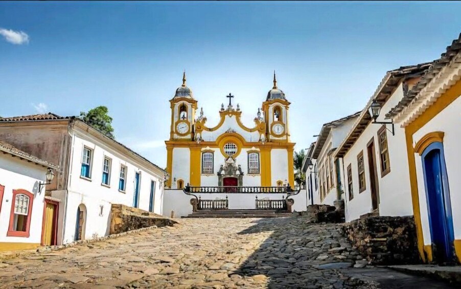 A cobblestone street leading to a yellow and white church with two bell towers. 1