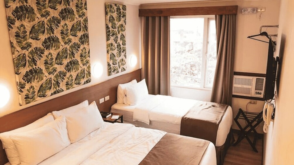 A hotel room with two beds, a window with curtains, a wall with leaf-patterned wallpaper, and a small desk with a telephone. 3