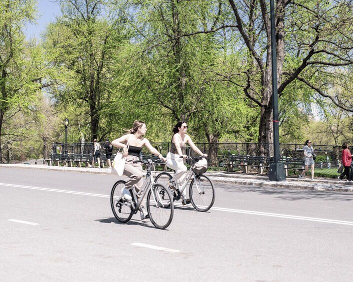 Two people riding bicycles on a road with trees and a fence in the background. 4