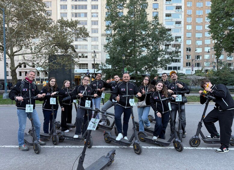 A group of people on electric scooters in an urban setting with buildings and trees in the background. 1