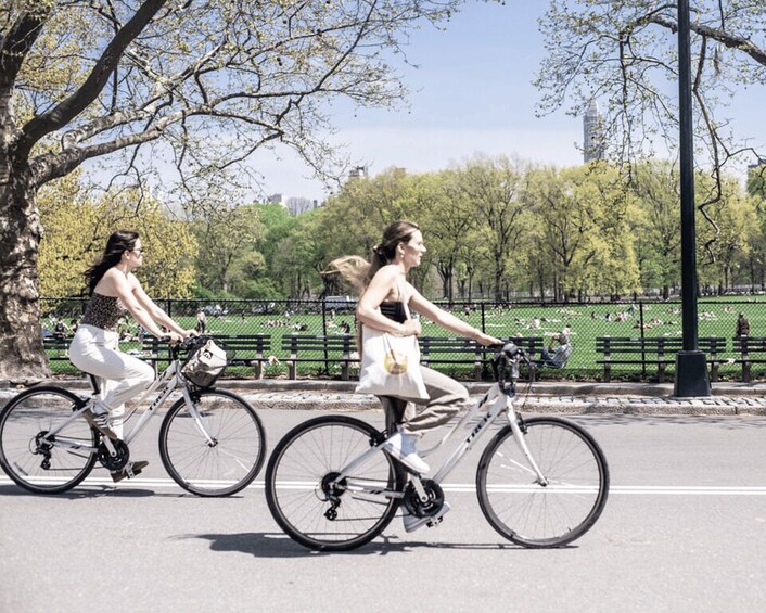 Two people riding bicycles on a park path with trees and a fence in the background. 3