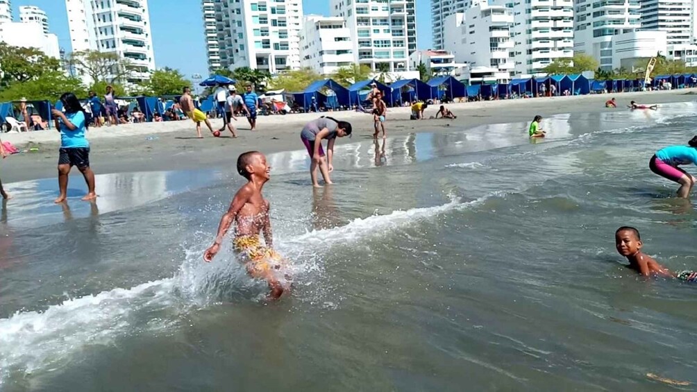 Children playing in shallow water at a beach with tents and buildings in the background. 5