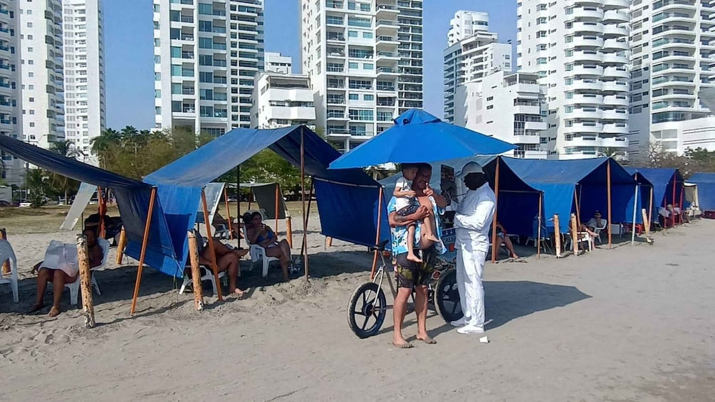 A beach with people under blue umbrellas, a man in white, and tall buildings in the background. 4