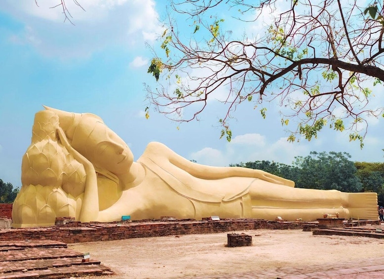 A large reclining Buddha statue in an open area with brick structures and a tree with sparse leaves. 2