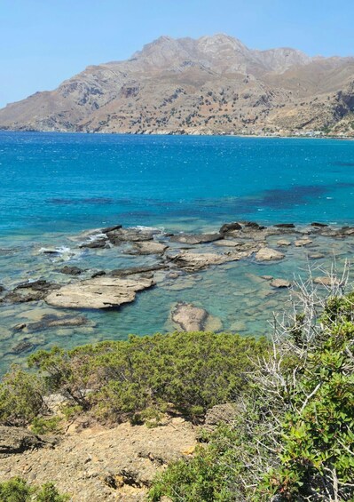 A coastal landscape with clear blue sea, rocky shore, and mountains in the background. 2