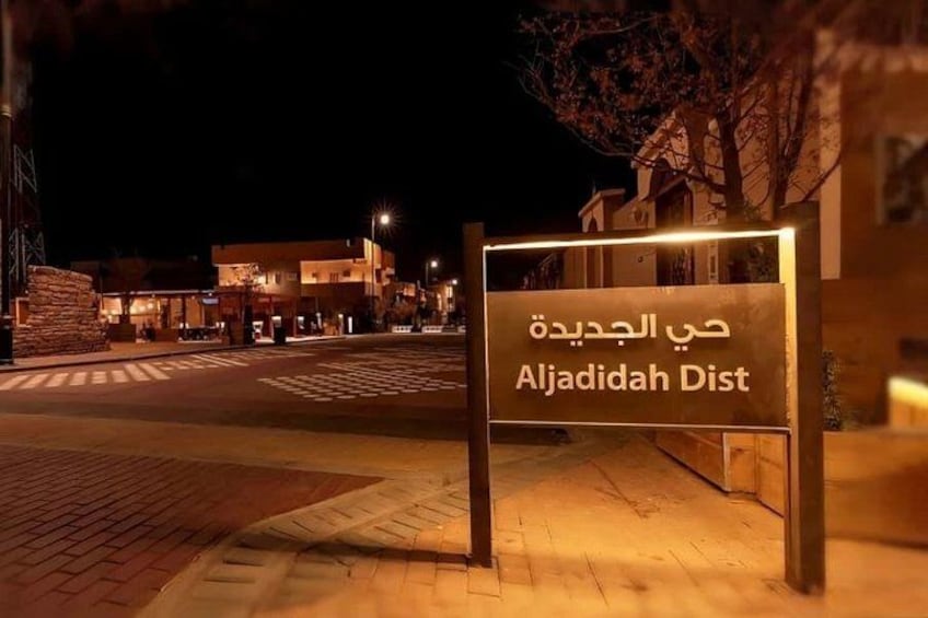 A nighttime street scene with a sign reading 'Aljadhidah Dist' in Arabic and English. 2