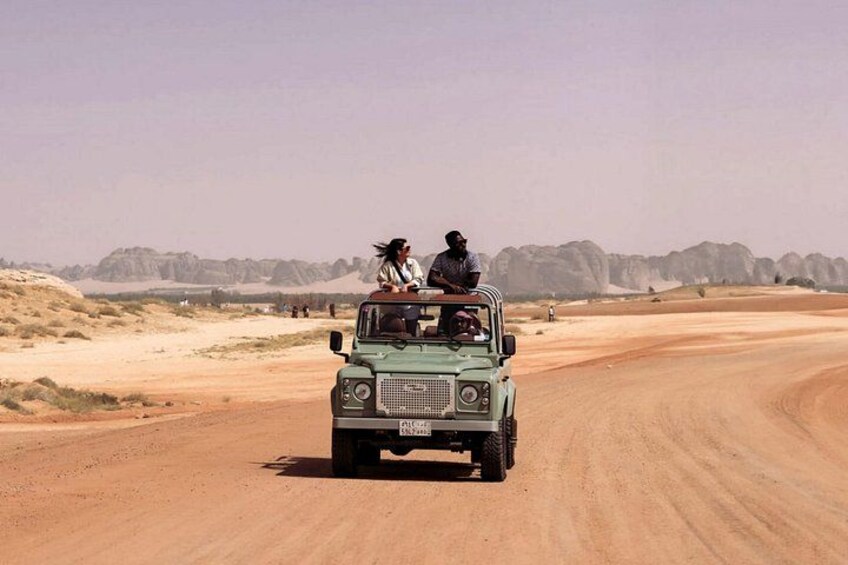 A green off-road vehicle driving on a dirt road in a desert landscape with rocky hills in the background. 5