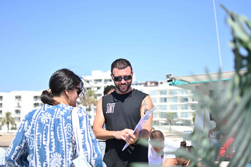 A person in a black sleeveless shirt with 'STAFF' written on it, holding a clipboard, standing near a pool with people in the background. 5