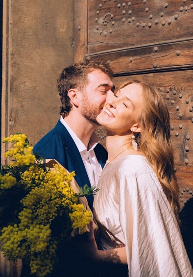 A couple embracing with a bouquet of yellow flowers in front of a wooden wall with riveted details. 4