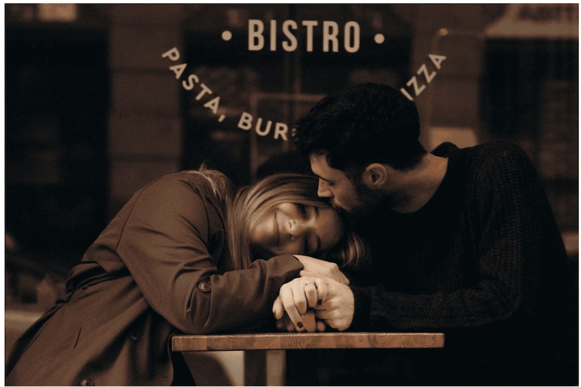 Two people leaning on a table in front of a bistro with signs for pasta, burgers, and pizza. 1