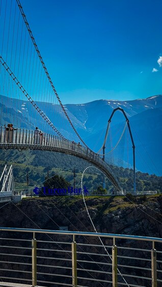 Een hangbrug met een metalen hek, een helderblauwe lucht en een bergachtig landschap op de achtergrond. 2