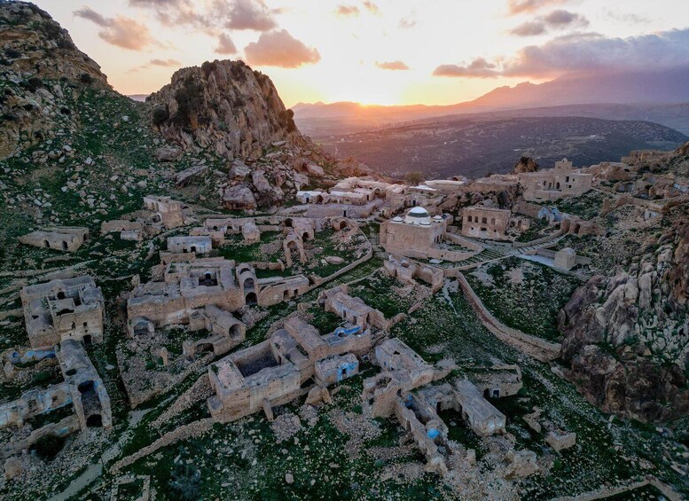 An ancient village with stone buildings and ruins, surrounded by rocky terrain and mountains, during sunset. 1