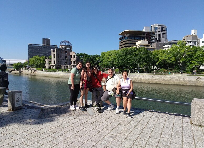 A group of people posing by a riverside walkway with modern buildings and trees in the background. 2