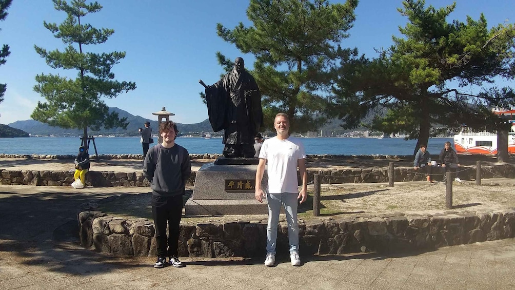 Two people standing in front of a statue with a body of water and trees in the background. 2
