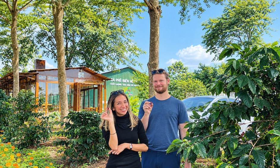 Two people standing in a garden with coffee plants and a building with a sign in the background. 4