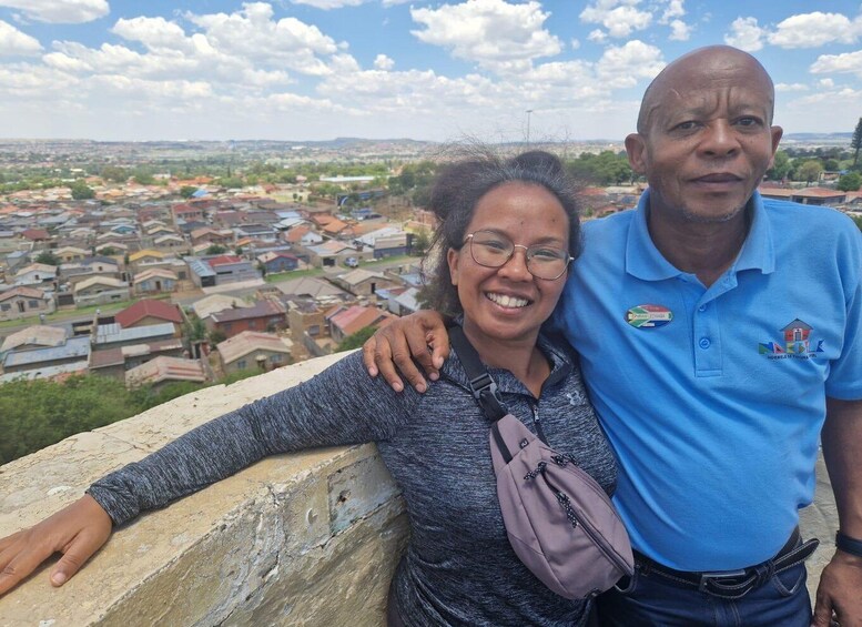 A man and a woman standing on a stone wall overlooking a densely packed residential area with various buildings and a clear sky. 1