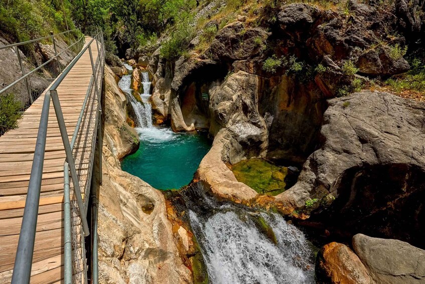 A wooden bridge over a rocky gorge with a turquoise pool and cascading waterfalls. 3