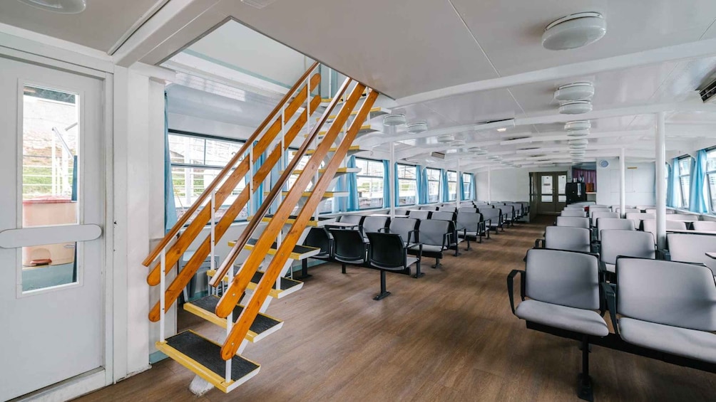 A ship's interior with a wooden staircase, rows of gray chairs, and large windows. 5