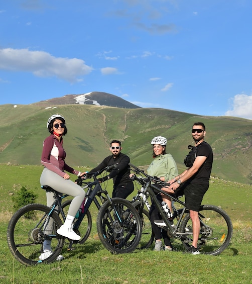 A group of people with bicycles on a grassy hill with a mountain in the background. 1