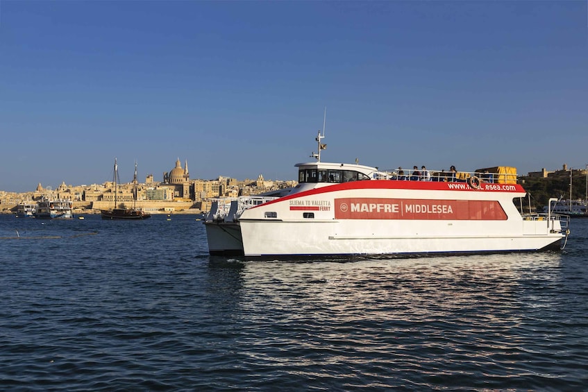 A white ferry named 'Mapfrel' with 'Sliena to Valletta Ferry' written on its side, sailing on the sea with a cityscape in the background. 2