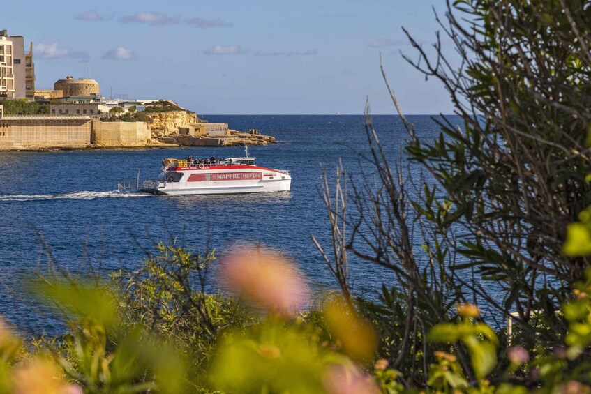 A white boat with red accents sailing on a calm sea, with buildings and a rocky coastline in the background. 1