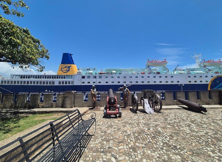 A cruise ship docked at a port with a soldier in historical uniform, cannons, and a bench. 4