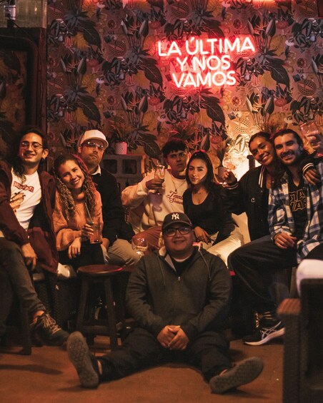 A group of people sitting and standing in a dimly lit room with a neon sign that reads 'LA ULTIMA Y NOS VAMOS'. 3