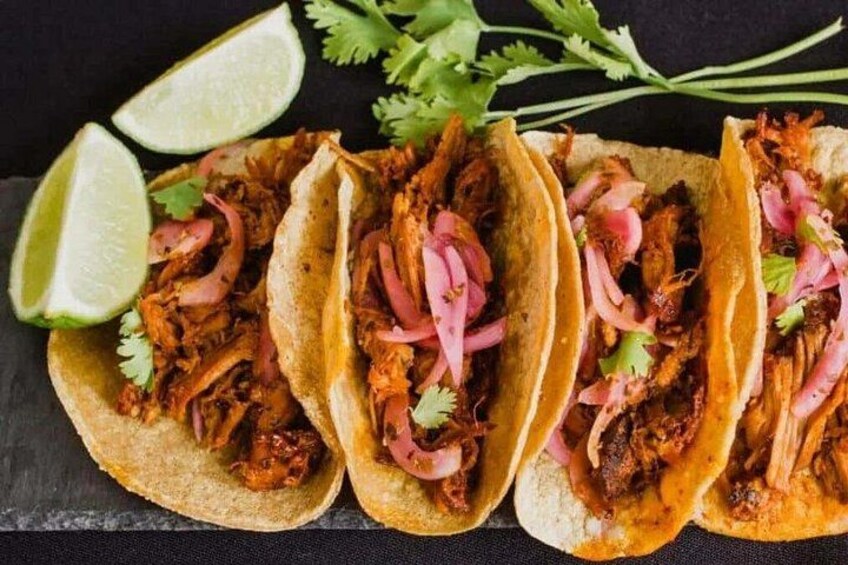 Three tacos with shredded meat, red onions, and cilantro, garnished with lime wedges on a dark surface. 2