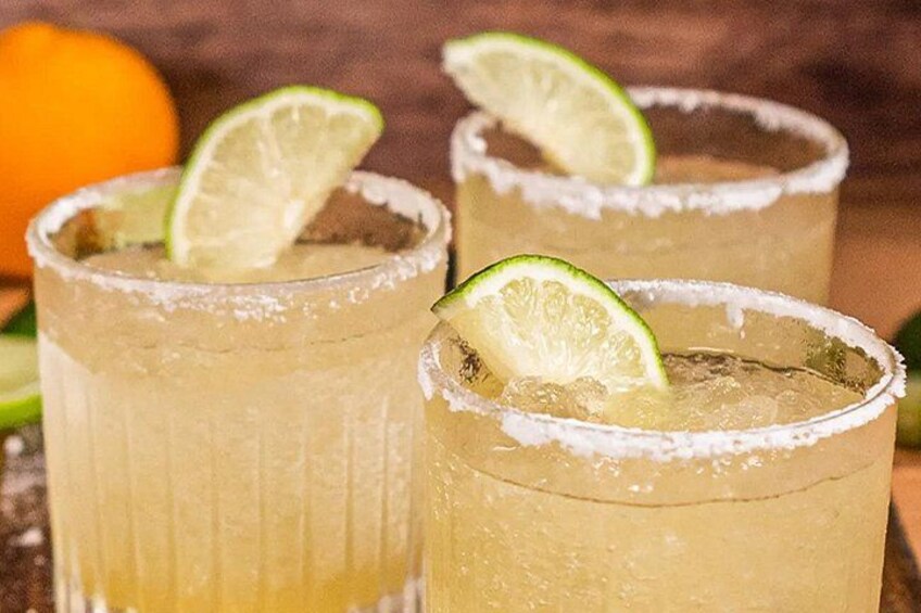 Three glasses of a yellow beverage with lime slices and salt on the rim, with an orange in the background. 2