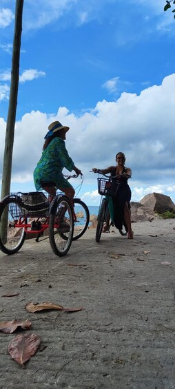 Two people riding bicycles on a dirt path with a clear sky and a large rock formation in the background. 3