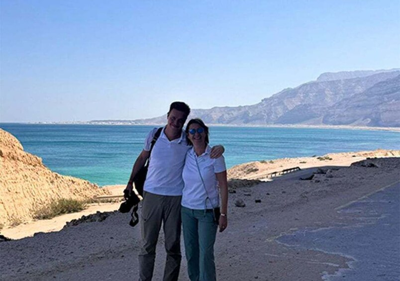 Två personer står på en strand med en klar himmel och en vattenyta i bakgrunden. 4