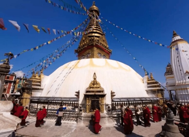 A large white stupa with golden spires, surrounded by people in red robes, and colorful prayer flags. 1