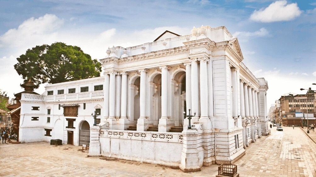 A classical white building with columns and a dome, surrounded by a cobblestone square and trees. 3