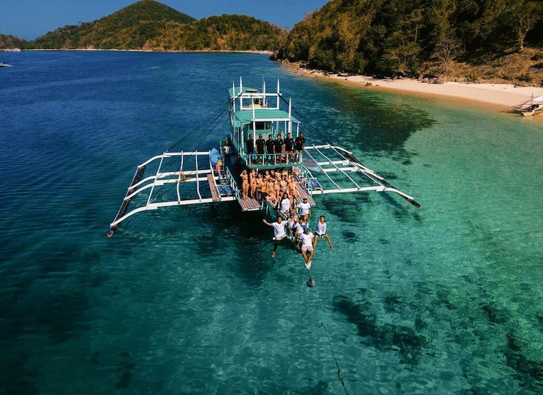 A boat with passengers in clear blue waters, a beach, and a forested hill in the background. 1