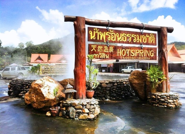 A sign for HOTSPRING THAWEESIN CHIANGRAJ with a traditional wooden archway, rocks, and potted plants. 2