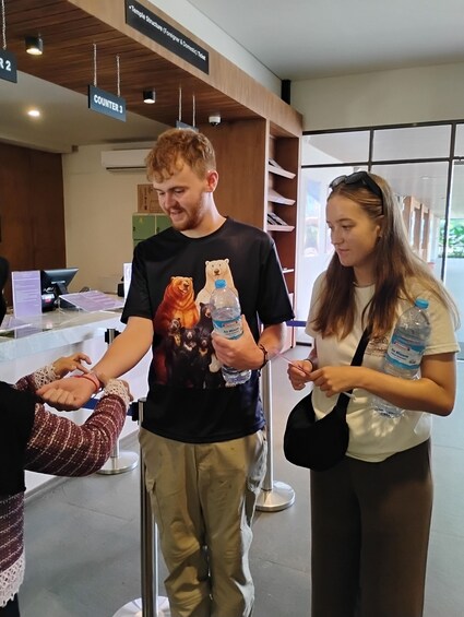 Henkilö, jolla on kädessään vesipullo ja lippu, seisoo rakennuksen palvelutiskillä. Rakennuksessa on puukatto ja opasteet, jotka osoittavat eri tiskien sijainnin. 3