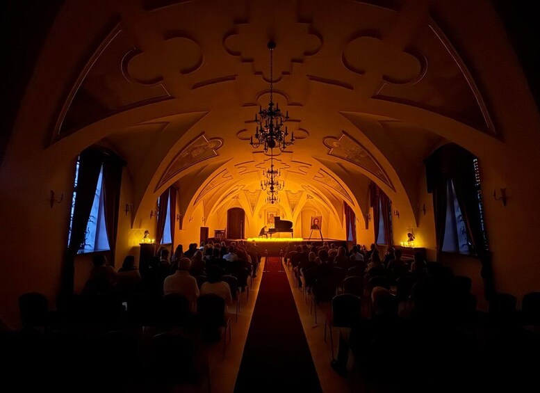An indoor concert hall with a grand piano on stage, an audience seated in rows, and a chandelier hanging from the ceiling. 2
