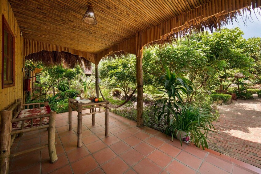 A covered outdoor area with a wooden table, chairs, and a thatched roof. 2