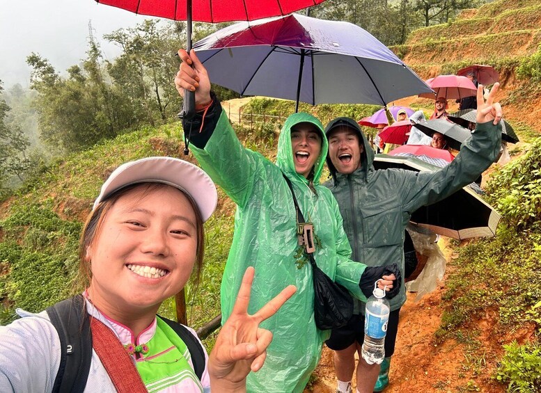 Three people hiking on a trail with rain gear, holding umbrellas, and one person making a peace sign. 1