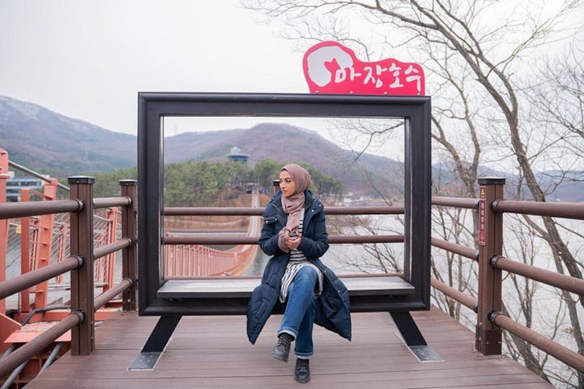 A person sitting on a wooden platform with a metal railing, overlooking a body of water and hills. 4