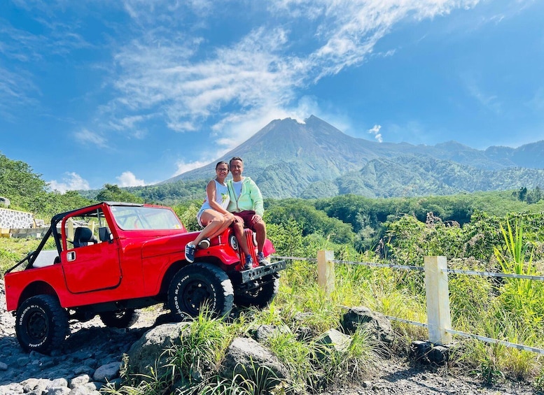 A red off-road vehicle with two people sitting on top, parked on a rocky path with a mountainous landscape in the background. 1