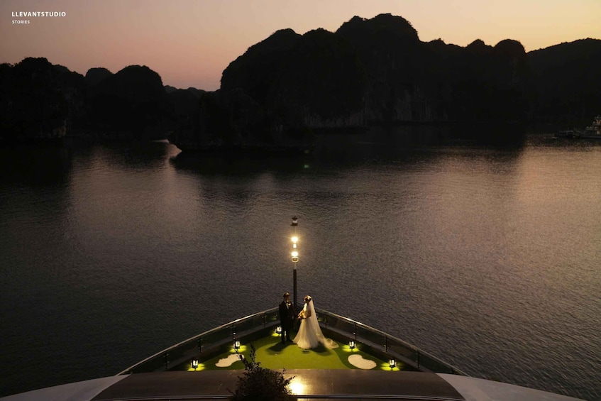 A boat with a couple standing at the bow, a calm body of water, and a mountainous landscape in the background. 2