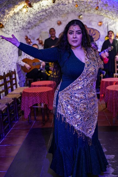 Una persona con un vestido azul oscuro y un chaleco estampado, de pie en una habitación con mesas cubiertas con manteles a cuadros rojos y blancos. 4