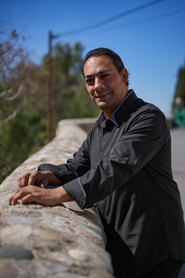 A man leaning on a stone wall, dressed in a dark shirt and tie, with a clear blue sky and greenery in the background. 3